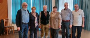 Sechs Personen stehen in einem Raum mit Holzboden und blauen Vorhängen. Eine Person trägt eine traditionelle Weste und braune Hose.