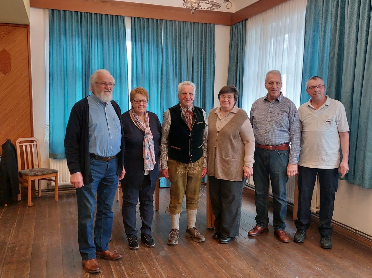 Eine Gruppe von sechs Personen, einschließlich Männer und Frauen, steht in einem Raum mit Holzböden. Sie alle lächeln und sehen in die Kamera.