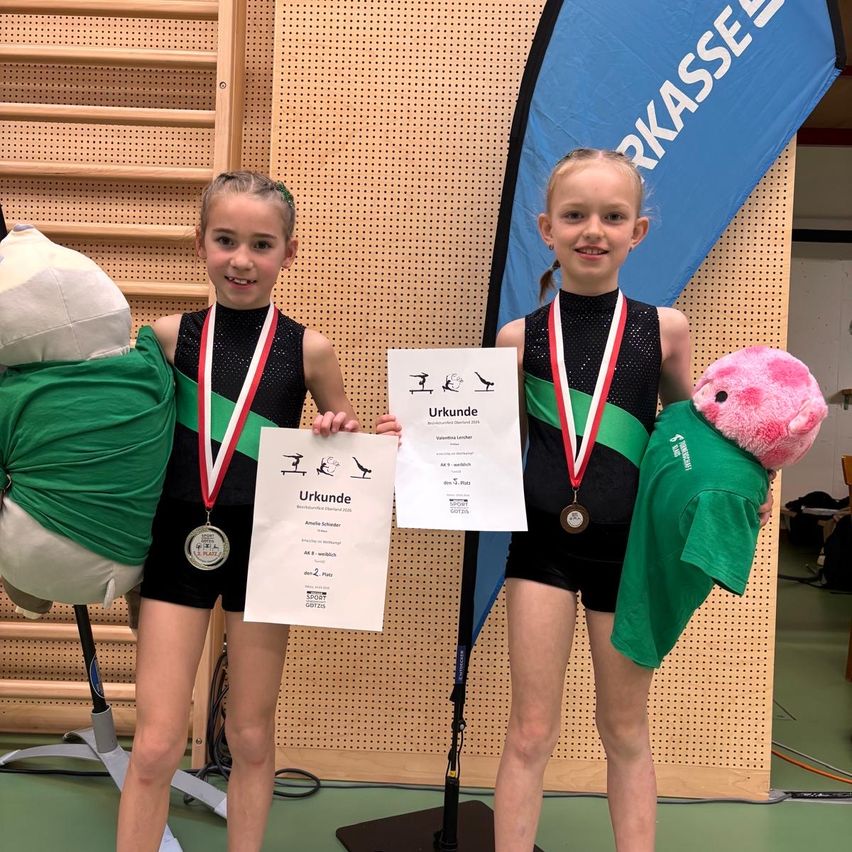 Zwei junge Mädchen mit Medaillen und Urkunden in einem Gymnastiksaal. Eine hält ein Kuscheltier und die andere eine Urkunde. Ein Banner mit dem Wort 'Kasse' ist hinter ihnen.