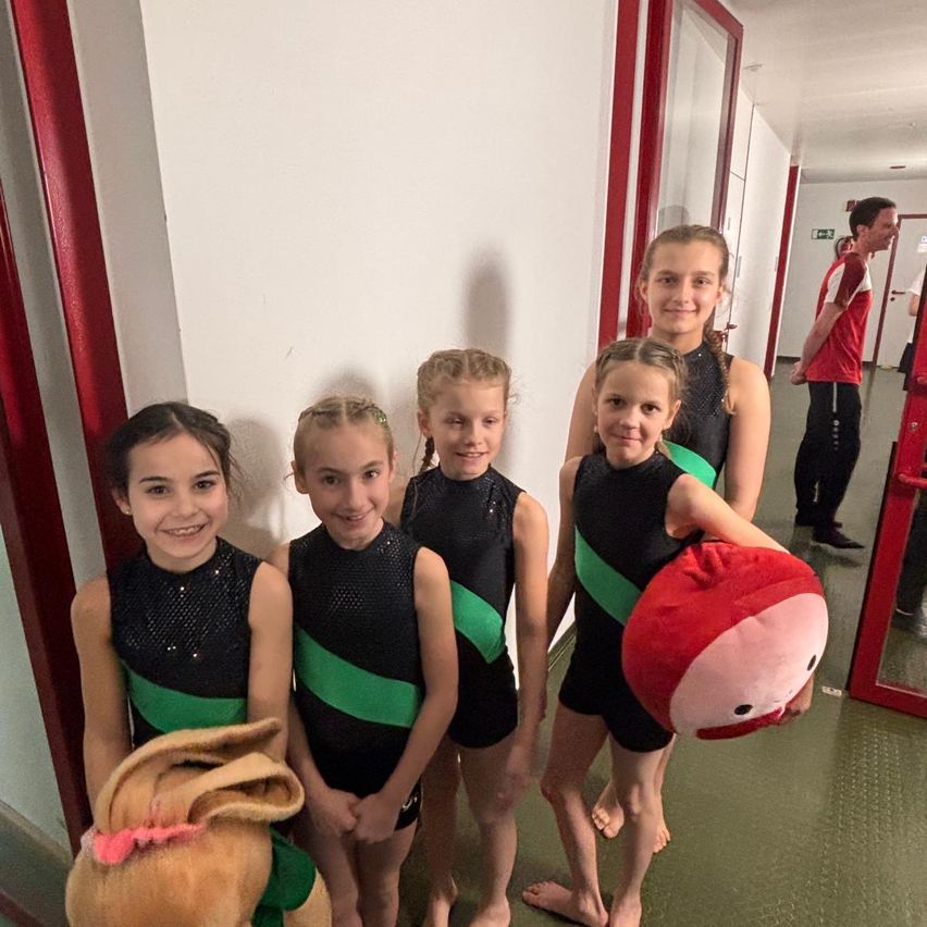 Fünf junge Mädchen in passenden schwarzen und grünen Outfits stehen lächelnd in einem Flur. Ein rotes Plüschtier wird von einem Mädchen gehalten. Ein anderes Mädchen hält ein braunes Plüschtier. Ein Mann steht in der Nähe einer Tür.