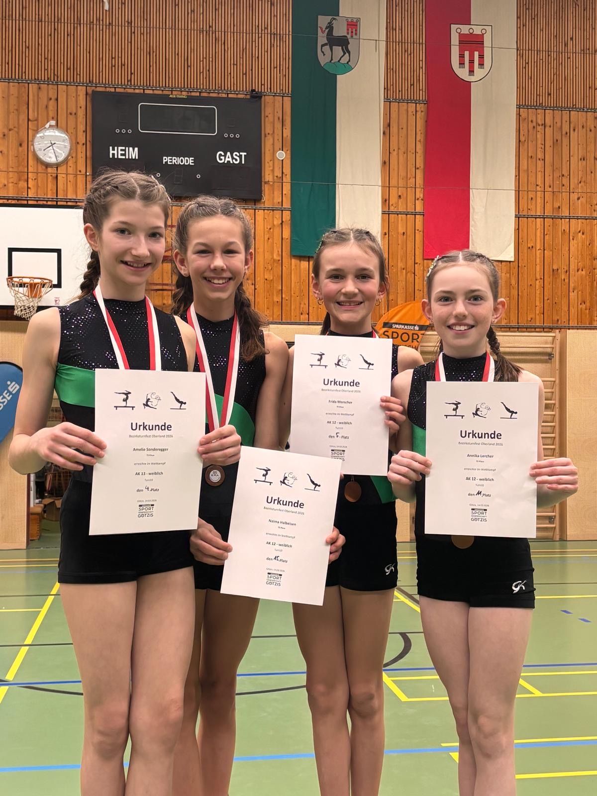 Vier Mädchen in Sportbekleidung posieren für ein Foto mit Urkunden in der Hand. Sie tragen Medaillen um den Hals. Hinter ihnen ist eine Turnhallenwand mit Flaggen und einem Basketballkorb.