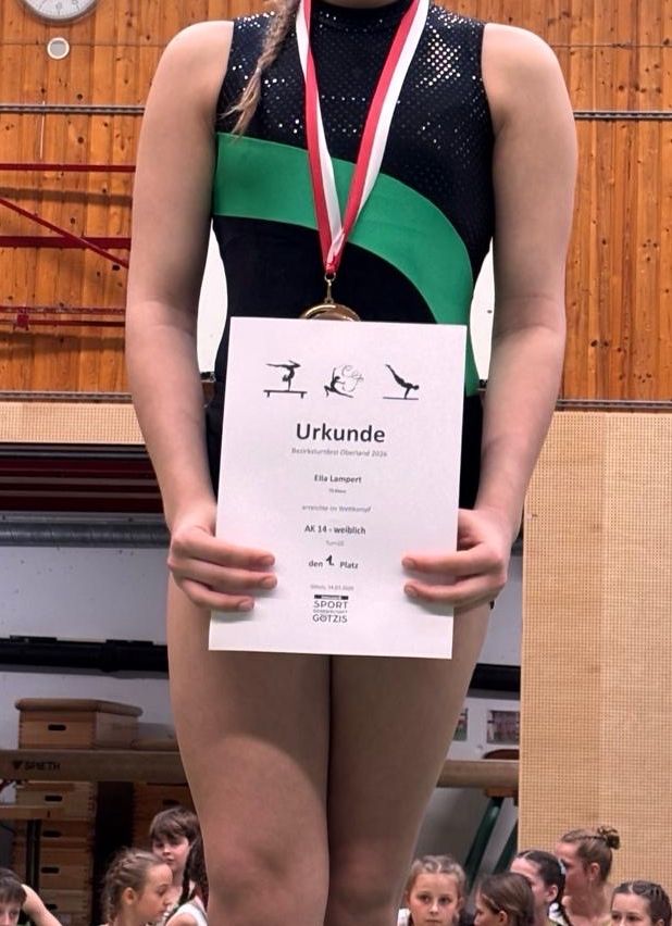 Ein Mädchen steht und hält eine Urkunde mit einer Medaille um den Hals. Die Urkunde trägt den Namen 'Ella Lampert'. Die Medaille ist gold mit grün-weißen Bändern. Der Hintergrund zeigt Holzwände.