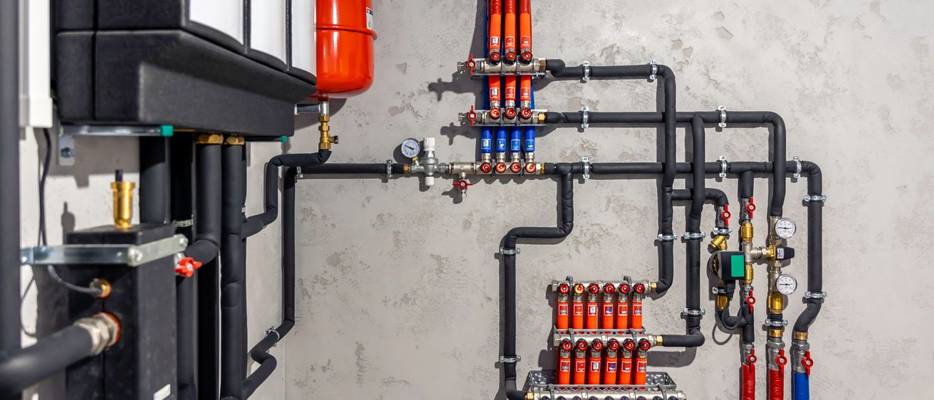 Eine Nahaufnahme einer Wand mit einem montierten System verschiedener Rohre und Armaturen, einschließlich eines roten Wassertanks und schwarzer Rohre mit blauen und roten Anschlüssen.