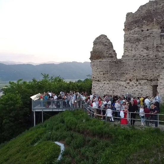 Eine Menschenmenge versammelt sich vor einer Burgruine auf einem Hügel, mit Blick auf Berge und Grün. Die Leute stehen und unterhalten sich, einige in der Nähe eines Tisches.