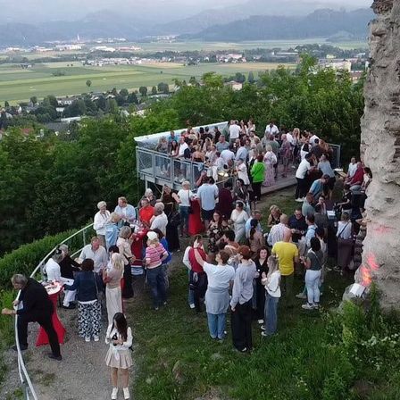 Eine Gruppe von Menschen, vermutlich auf einem Dach, versammelt sich in der Nähe eines Zauns. Sie stehen um Tische herum, unterhalten sich und einige sitzen. Im Hintergrund sind Bäume und Gebäude zu sehen.