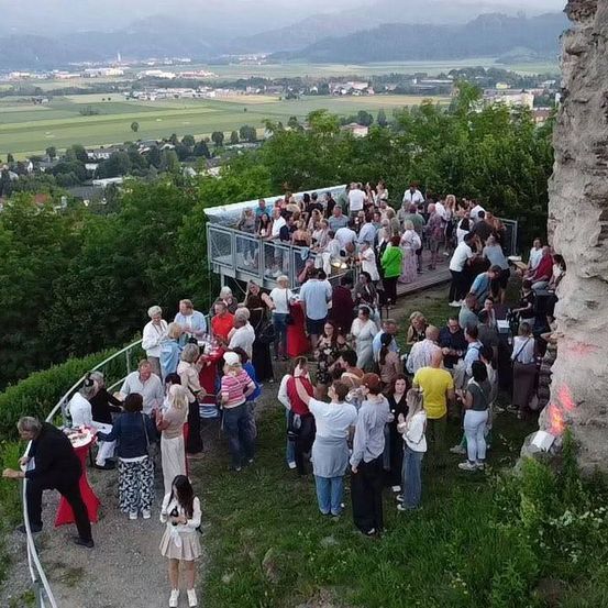 Eine Gruppe von Menschen, vermutlich auf einem Dach, versammelt sich in der Nähe eines Zauns. Sie stehen um Tische herum, unterhalten sich und einige sitzen. Im Hintergrund sind Bäume und Gebäude zu sehen.