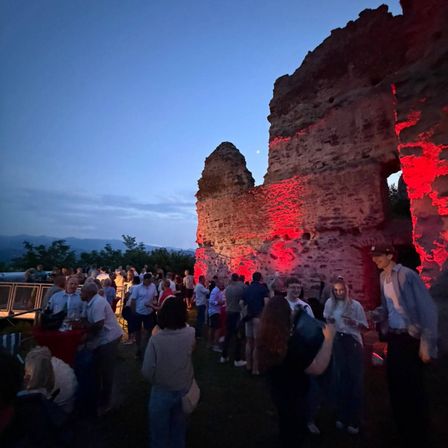 Eine Menschenmenge steht vor einer von roten Lichtern erleuchteten historischen Ruine in der Dämmerung, mit Bergen im Hintergrund.