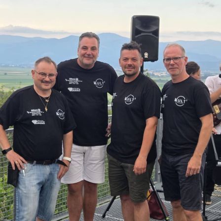Vier Männer in passenden schwarzen T-Shirts mit weißen Logos stehen auf einer Brücke und lächeln für ein Foto vor einem bergigen Hintergrund mit einem Lautsprecher dahinter.