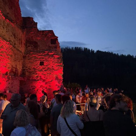 Menschenmengen versammeln sich bei roter Beleuchtung in der Nacht, vor einer erleuchteten alten Steinstruktur, mit Bäumen im Hintergrund.