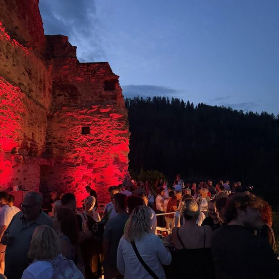 Menschenmengen versammeln sich bei roter Beleuchtung in der Nacht, vor einer erleuchteten alten Steinstruktur, mit Bäumen im Hintergrund.