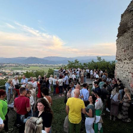 Eine große Menschenmenge versammelt sich auf einer grasbewachsenen Anhöhe mit einer Steinmauer und überblickt ein Tal mit Gebäuden und Bergen unter einem klaren, blauen Himmel.