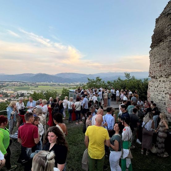 Eine große Menschenmenge versammelt sich auf einer grasbewachsenen Anhöhe mit einer Steinmauer und überblickt ein Tal mit Gebäuden und Bergen unter einem klaren, blauen Himmel.