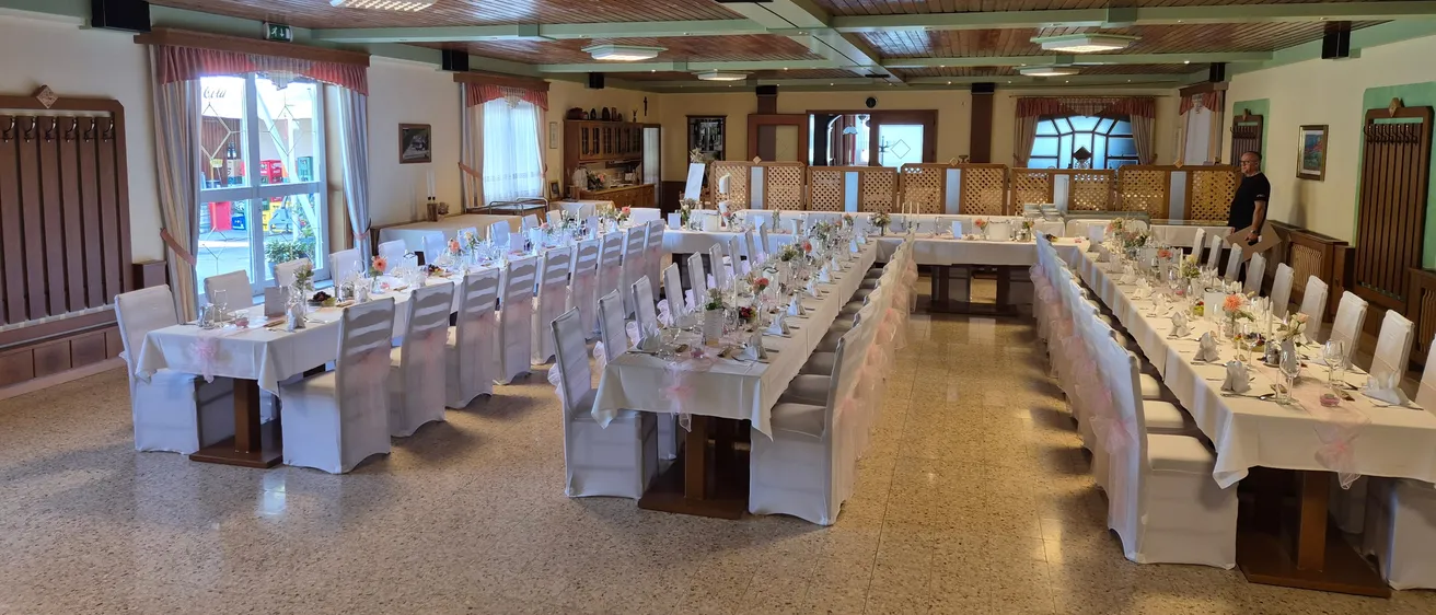 Ein großer Speisesaal mit mehreren langen Tischen, die für eine formelle Veranstaltung vorbereitet sind, geschmückt mit weißen Tischdecken, Blumengestecken und ordentlich angeordneten Stühlen. Holzbalken und Deckenleuchten tragen zur eleganten Atmosphäre bei.
