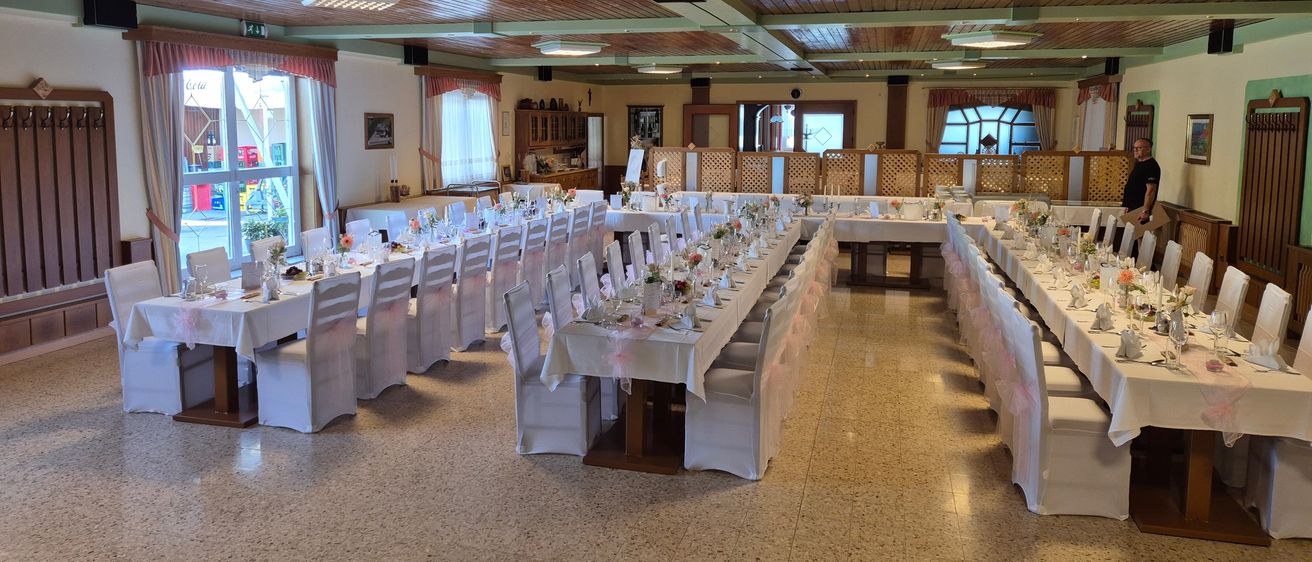 Ein großer Speisesaal mit mehreren langen Tischen, die für eine formelle Veranstaltung vorbereitet sind, geschmückt mit weißen Tischdecken, Blumengestecken und ordentlich angeordneten Stühlen. Holzbalken und Deckenleuchten tragen zur eleganten Atmosphäre bei.
