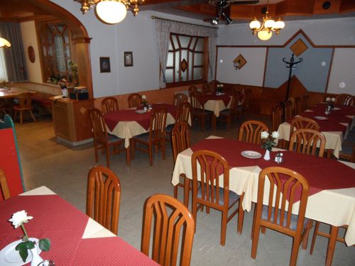 Ein Restaurantinnenraum mit Holzstühlen und -tischen, weißen Tischdecken und einem Deckenventilator. Auf einigen Tischen stehen Blumen in Vasen.