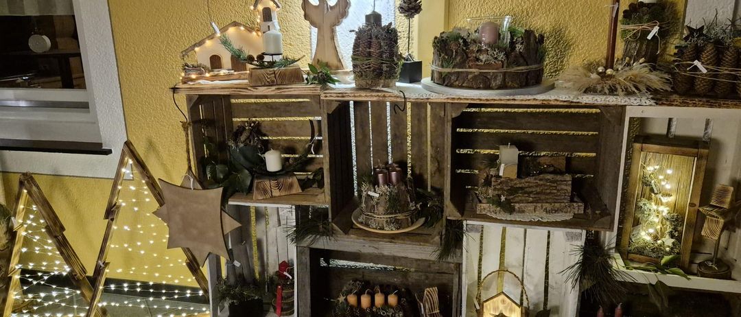 Ein Holzkistenregal zeigt festliche Gegenstände, darunter Kerzen, Pflanzen und Ornamente. Links stehen ein Stern und eine weihnachtlich beleuchtete Krippe, rechts eine Laterne und ein sternförmiges Objekt. Der Hintergrund zeigt eine gelbe Wand.