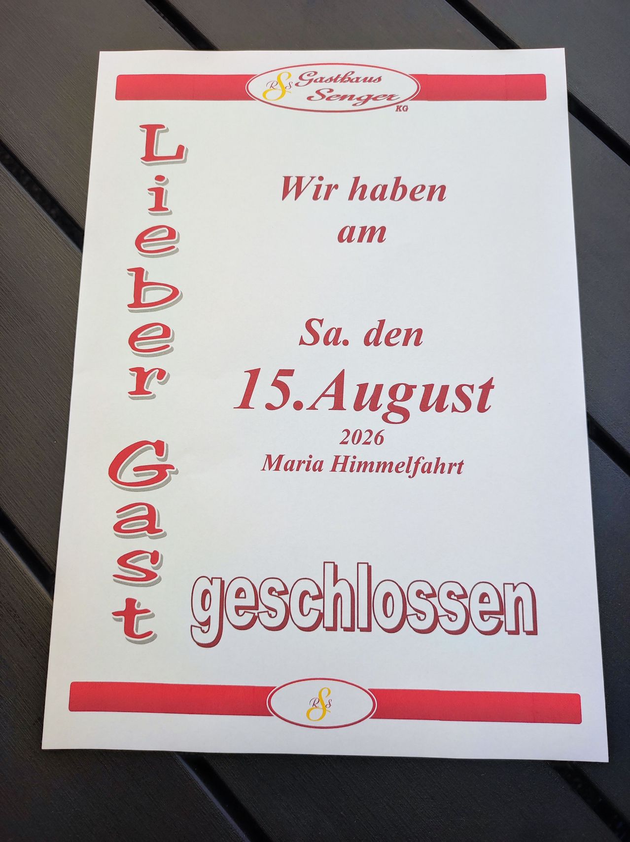 Ein weißes Schild mit rotem Text lautet 'Lieber Gast, Wir haben am Sa. den 15. August 2026 Maria Himmelfahrt geschlossen.' Es steht auf einer Holzoberfläche.