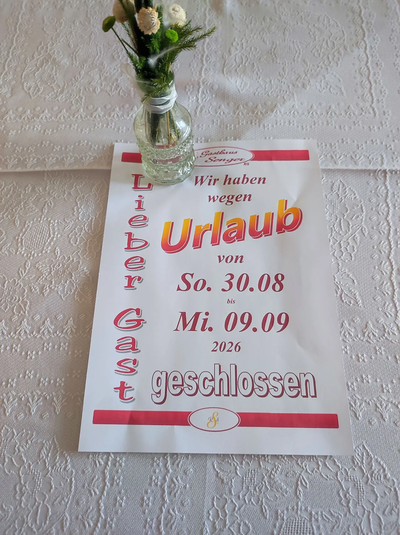Ein weißes Schild mit rotem Text steht 'Lieber Gast, wir haben vom 30.08.2026 bis 09.09.2026 geschlossen.' Ein kleines Glasvase mit Blumen steht auf dem Schild.