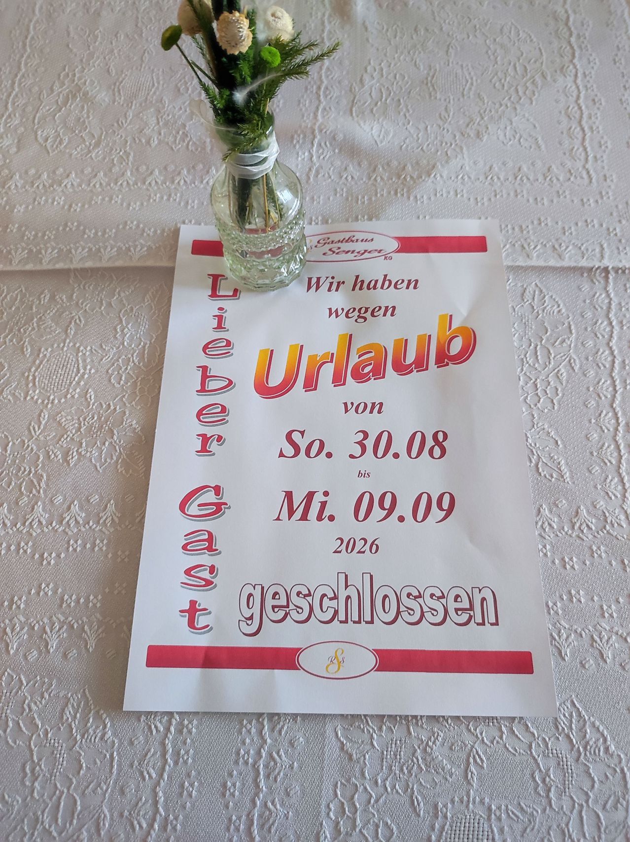 Ein weißes Schild mit rotem Text steht 'Lieber Gast, wir haben vom 30.08.2026 bis 09.09.2026 geschlossen.' Ein kleines Glasvase mit Blumen steht auf dem Schild.
