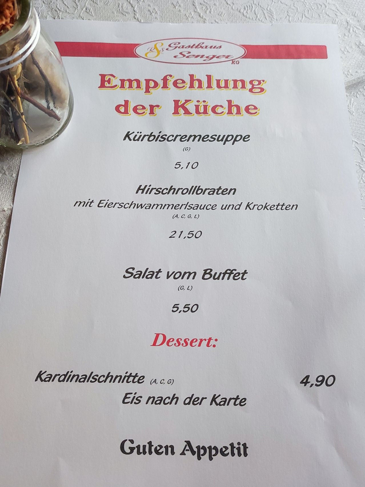 Ein Menü mit einem Glas im Hintergrund. Es bietet Gerichte wie 'Kürbiscremesuppe', 'Hirschroldbraten', 'Salat vom Buffet' und 'Dessert: Kardinalschnitte'. Die Preise sind neben jedem Gericht aufgeführt.