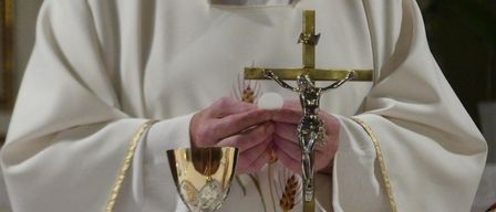 Ein Priester hält ein Kreuz und einen Kelch mit einem konsekrierten Hostien, gekleidet in einer weißen Tunika mit goldenem Rand.