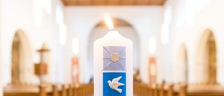 A candle with four panels on it is lit in a church. The candle is white and has a flame. The panels depict a dove, a fish, footprints, and a sun.