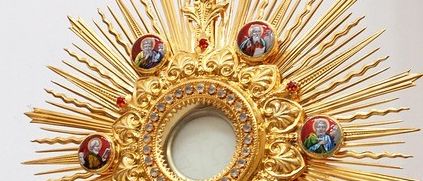 Ein goldener Monstranz mit einem Kreuz an der Spitze und einem runden Rahmen darum, geschmückt mit roten Edelsteinen und Bildern von Heiligen.
