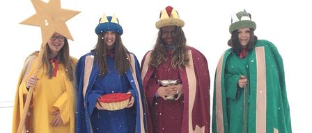 Four people dressed in festive costumes are standing in the snow. They are wearing crowns, holding a basket, a cup, and a lantern. They seem to be part of a Christmas celebration.