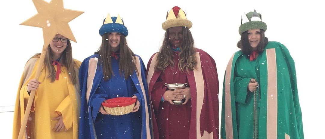 Vier Menschen in festlichen Kostümen stehen im Schnee. Sie tragen Kronen und halten einen Korb, einen Becher und eine Laterne. Sie scheinen Teil einer Weihnachtsfeier zu sein.