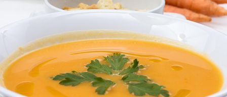 Eine Schüssel mit orangefarbener Karottensuppe, garniert mit Petersilie. Im Hintergrund sind frische Karotten zu sehen.
