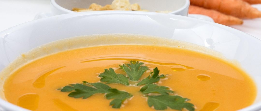 Eine Schüssel mit orangefarbener Karottensuppe, garniert mit Petersilie. Im Hintergrund sind frische Karotten zu sehen.