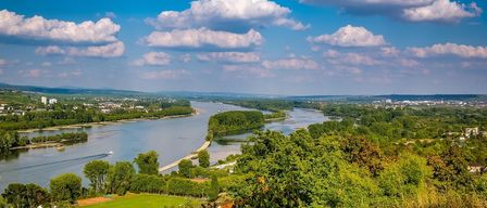 Luftaufnahme eines Flusses, der durch ein Tal mit üppiger Vegetation fließt, einer Stadtsilhouette in der Ferne und einem klaren blauen Himmel mit verstreuten Wolken.