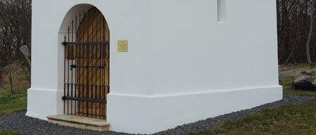 Eine kleine weiße Kapelle mit grauem Dach, einem Kreuz obenauf und einem hölzernen Tor steht auf einem Grasfeld mit Bäumen im Hintergrund.