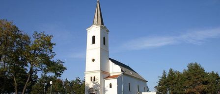 A white church with a steeple stands on a hill surrounded by green grass and trees under a blue sky.