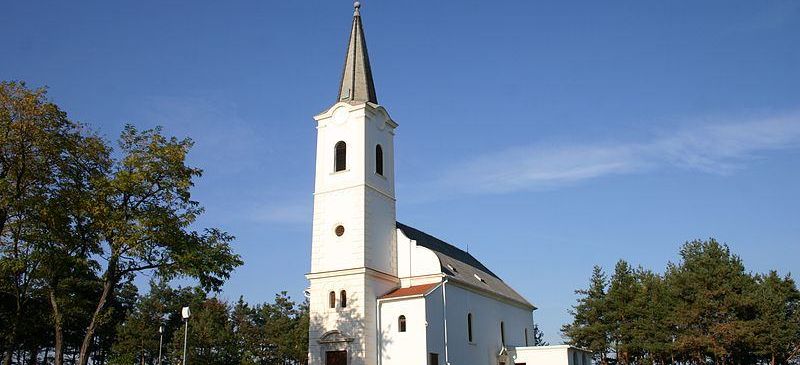 Eine weiße Kirche mit einem Turm steht auf einem Hügel, umgeben von grünem Gras und Bäumen unter einem blauen Himmel.