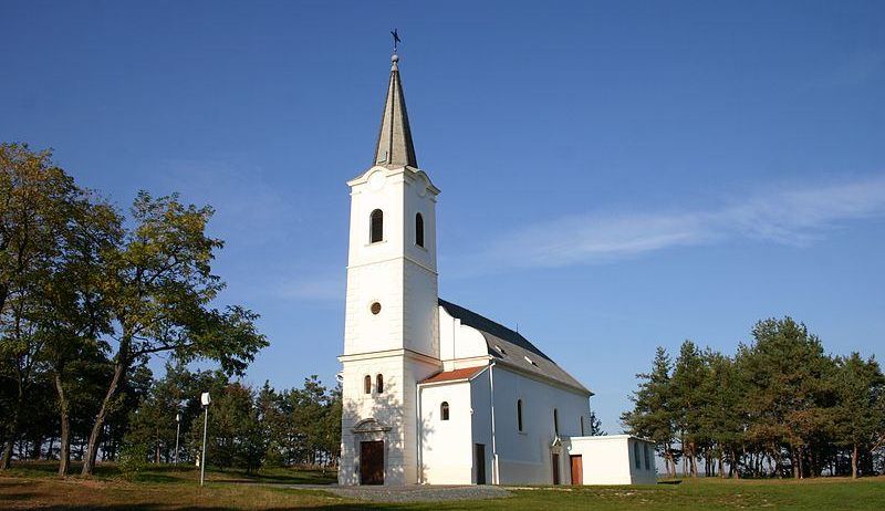 Eine weiße Kirche mit einem Turm steht auf einem Hügel, umgeben von grünem Gras und Bäumen unter einem blauen Himmel.