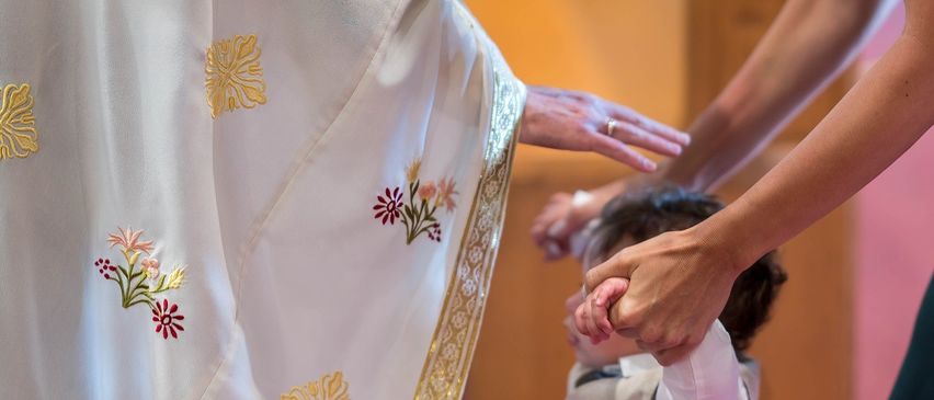 Ein Priester hält die Hand eines Kindes während einer religiösen Zeremonie, gekleidet in einen weißen Gewand mit goldenen Blumendesigns.