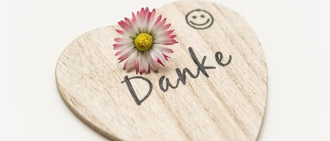 Ein herzförmiges Holzobjekt mit einer Blume und dem Wort 'Danke' darauf. Die Blume ist rosa mit einem gelben Zentrum. Das Wort 'Danke' ist in Schwarz geschrieben. Der Hintergrund ist weiß.