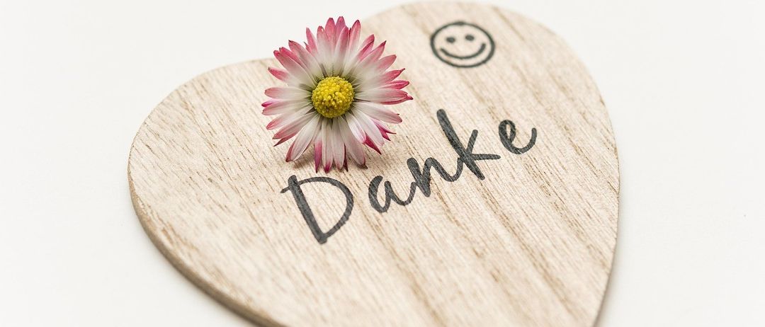 Ein herzförmiges Holzobjekt mit einer Blume und dem Wort 'Danke' darauf. Die Blume ist rosa mit einem gelben Zentrum. Das Wort 'Danke' ist in Schwarz geschrieben. Der Hintergrund ist weiß.