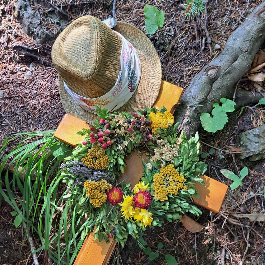 Ein Strohhut liegt auf einem Holzkreuz, das mit einem Kranz aus gelben und roten Blumen geschmückt ist, umgeben von Grün, auf einem Waldboden.