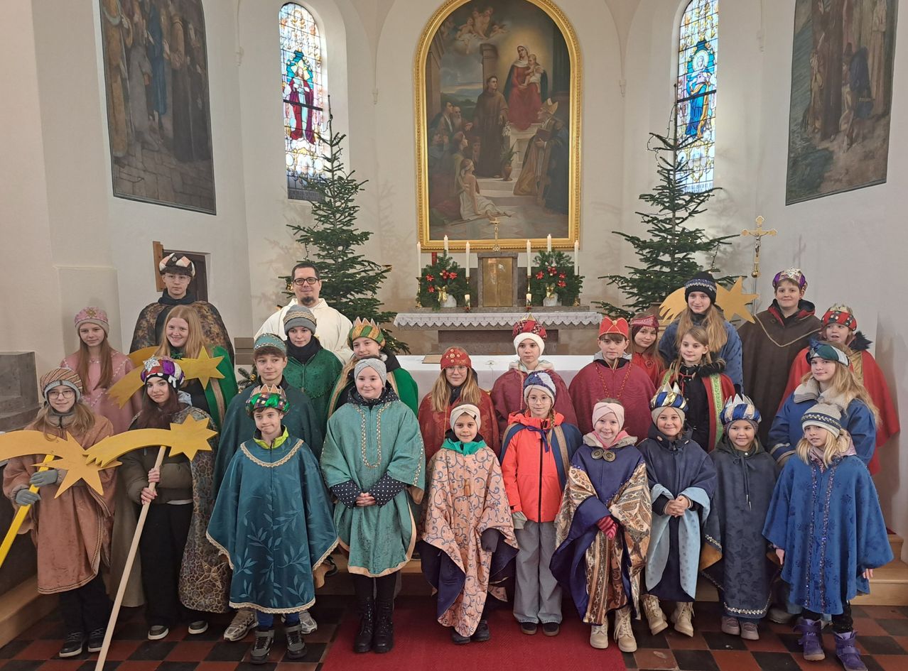 Eine Gruppe von Kindern in Kostümen posiert für ein Foto in einer Kirche mit Altar, Weihnachtsbäumen und Kerzen.
