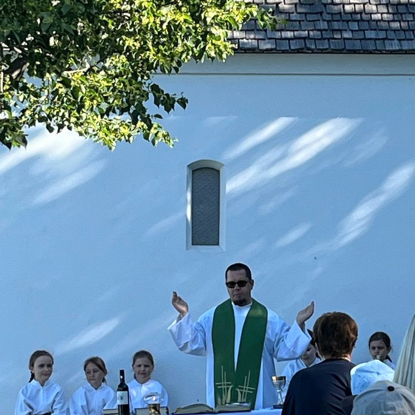 Ein Priester leitet einen Gottesdienst unter einem Baum vor einer Kirche, während eine Gruppe von Menschen zuhört. Flaschen und ein Weinglas stehen auf dem Tisch.