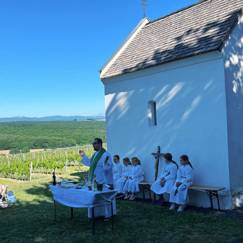 Ein Priester in einer weißen Robe spricht zu Kindern in Weiß, die vor einer kleinen weißen Kirche sitzen. Ein Tisch mit Weinflaschen und einem Kreuz steht vor ihnen.