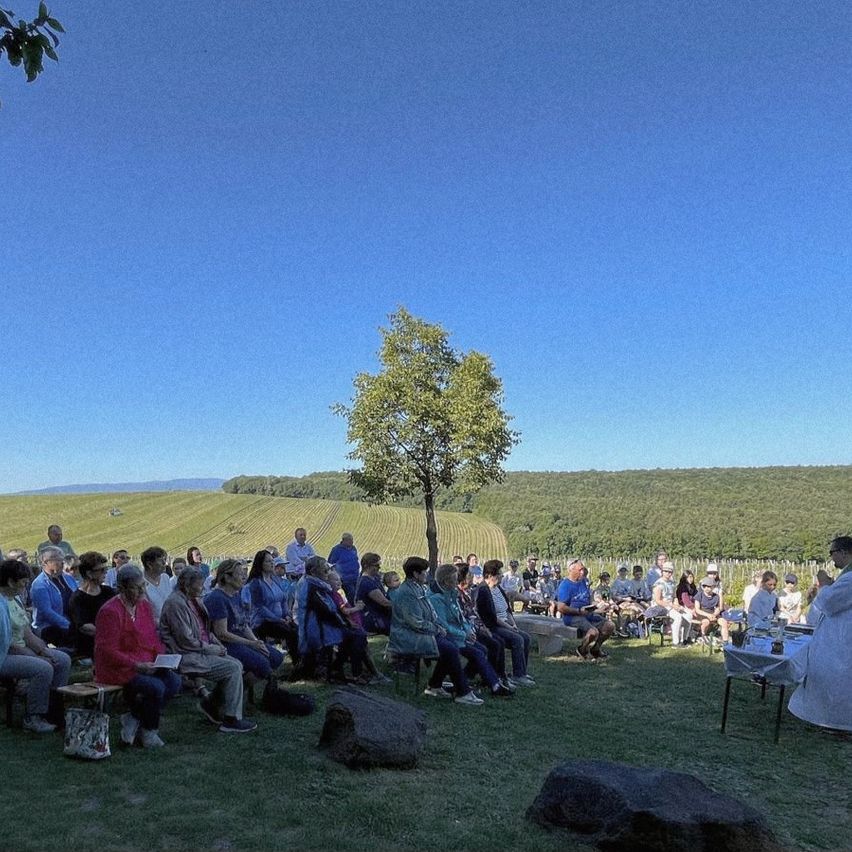 Eine Gruppe von Menschen hat sich an einem sonnigen Tag im Freien versammelt, sitzt auf Bänken und Stühlen. Ein Priester steht vor ihnen. Die Umgebung ist von Bäumen und Gras umgeben. In der Ferne gibt es ein Feld und eine Bergkette.