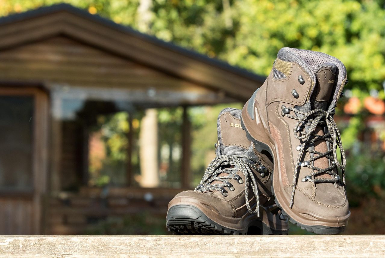 A pair of brown hiking boots with laces and metal buckles, resting on a wooden bench with a blurry background of a building and trees.