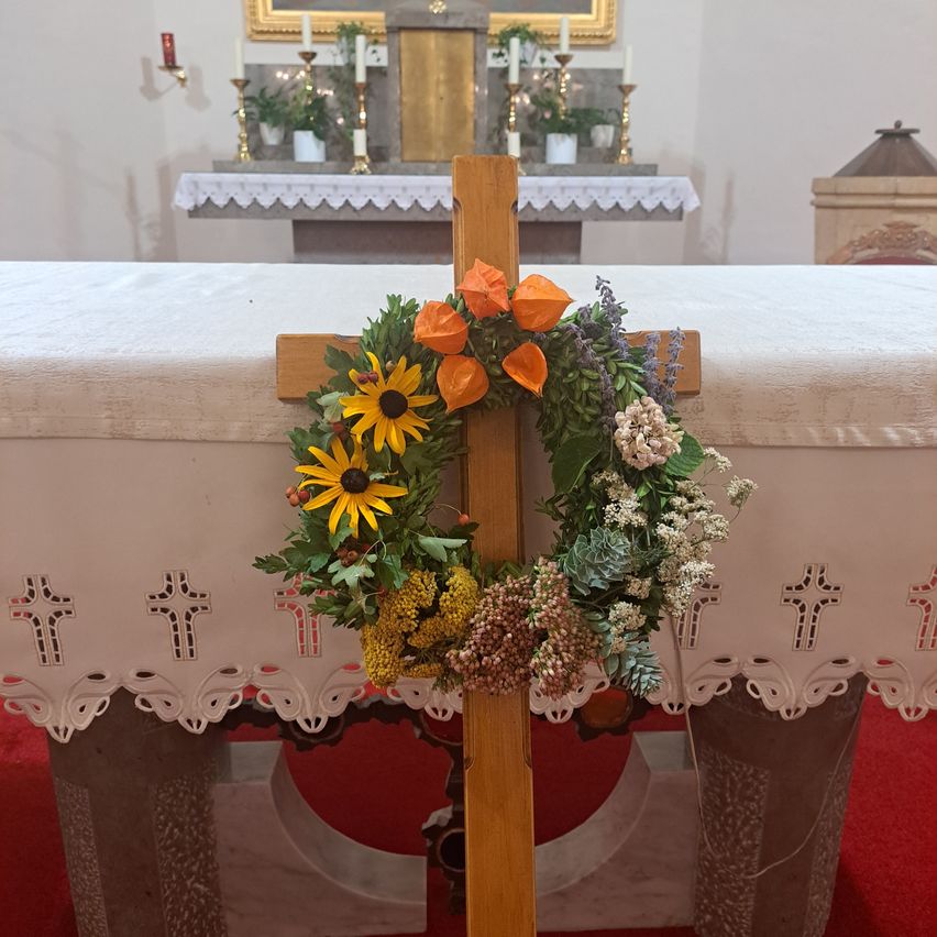 A wooden cross adorned with a wreath of sunflowers, orange flowers, and green leaves stands in front of a white altar in a church.