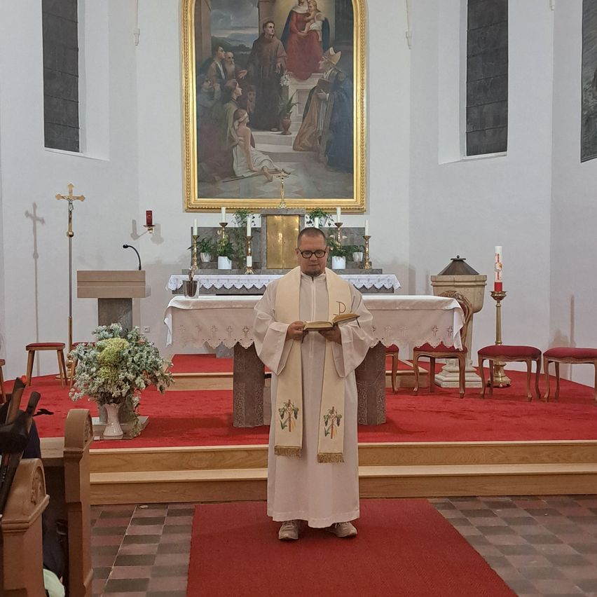 A priest stands in front of an altar in a church, holding a book. Behind him is a large painting on the wall.