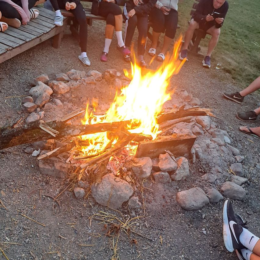 Eine Gruppe von Menschen sitzt um ein brennendes Feuer mit Steinen und Holz, das hell brennt, einige tragen Turnschuhe und Socken.