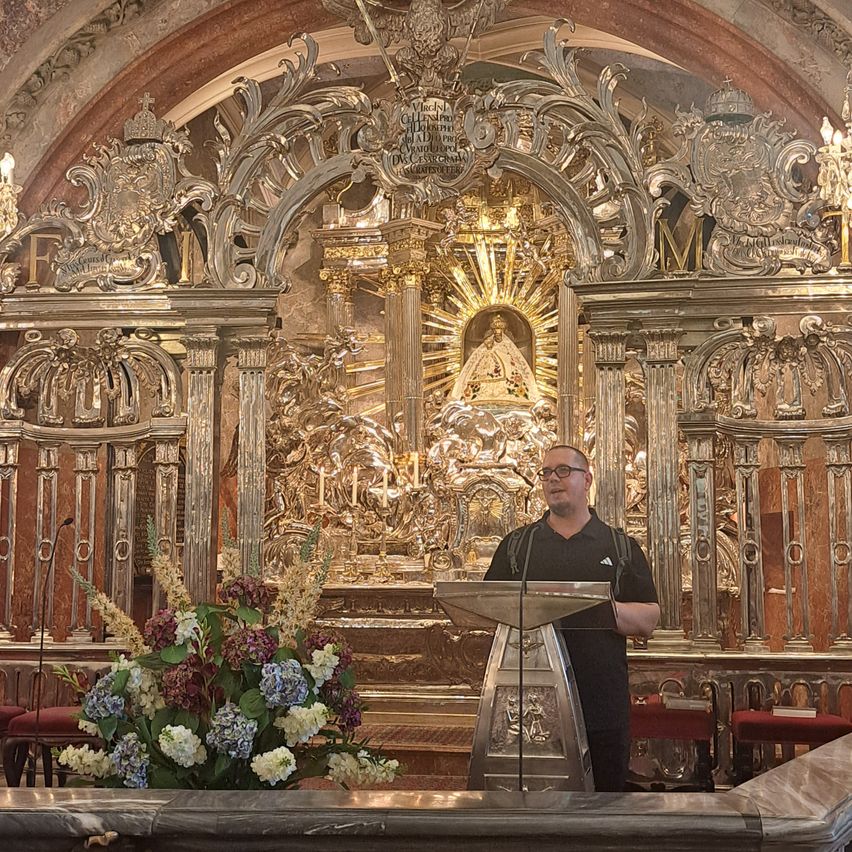 A man stands at a podium in a grand, ornate church with a golden altar and intricate decorations in the background.