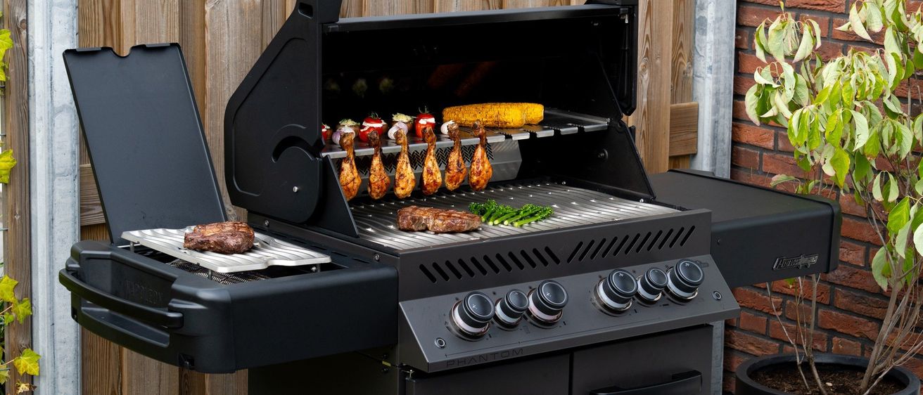 Ein schwarzer Outdoor-Grill brät verschiedene Fleischsorten und Gemüse auf einem Metallrost. In der Nähe steht ein Tablett mit weiterem Essen. Der Grill hat sechs Brenner und ein digitales Bedienfeld. Dahinter befindet sich ein Holzzaun.